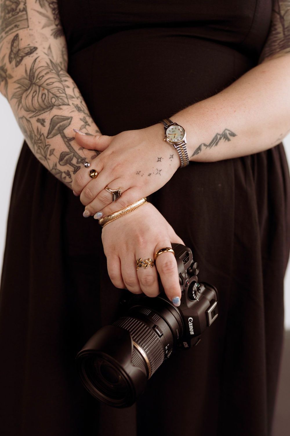 Close-up of Liv Finley's tattooed hands holding a Canon camera, gold rings and bracelets