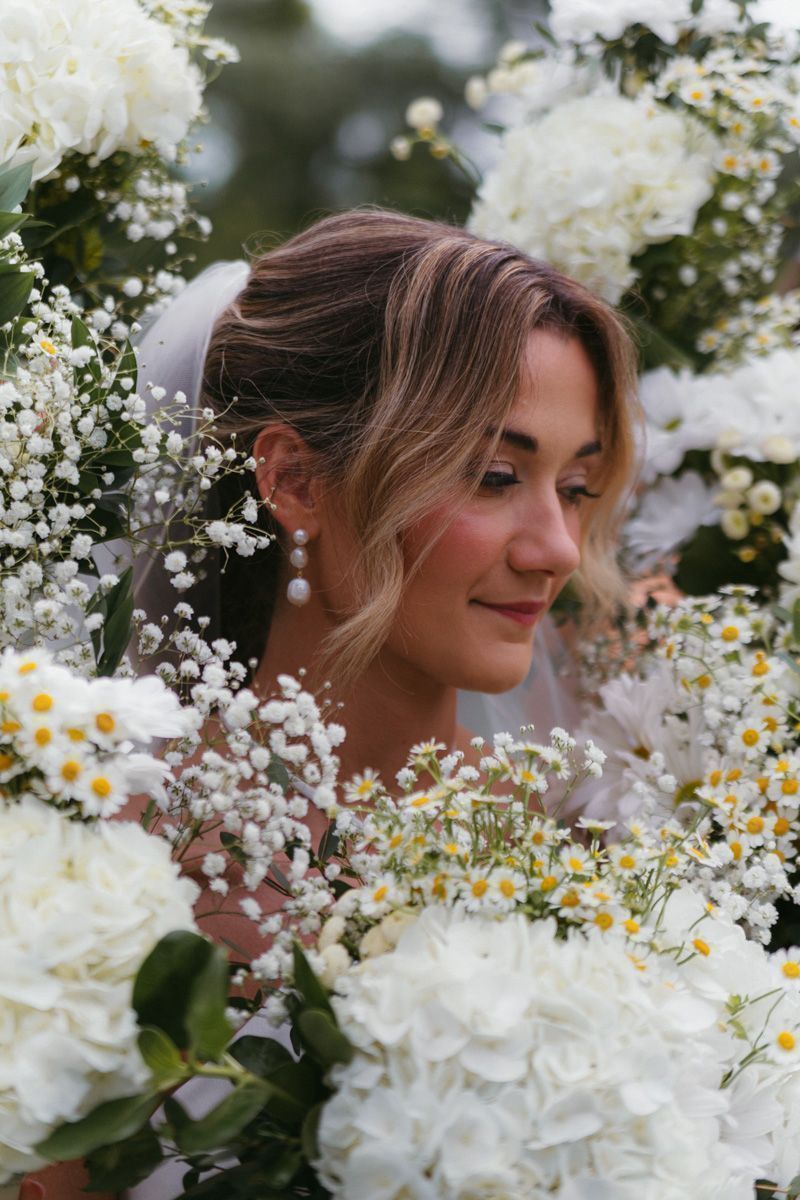 Bride gazing downward, framed by white hydrangeas and wildflowers, wearing a veil and pearl drop earrings