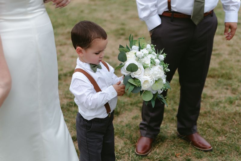Young ring bearer in white shirt and suspenders holding the bridal bouquet