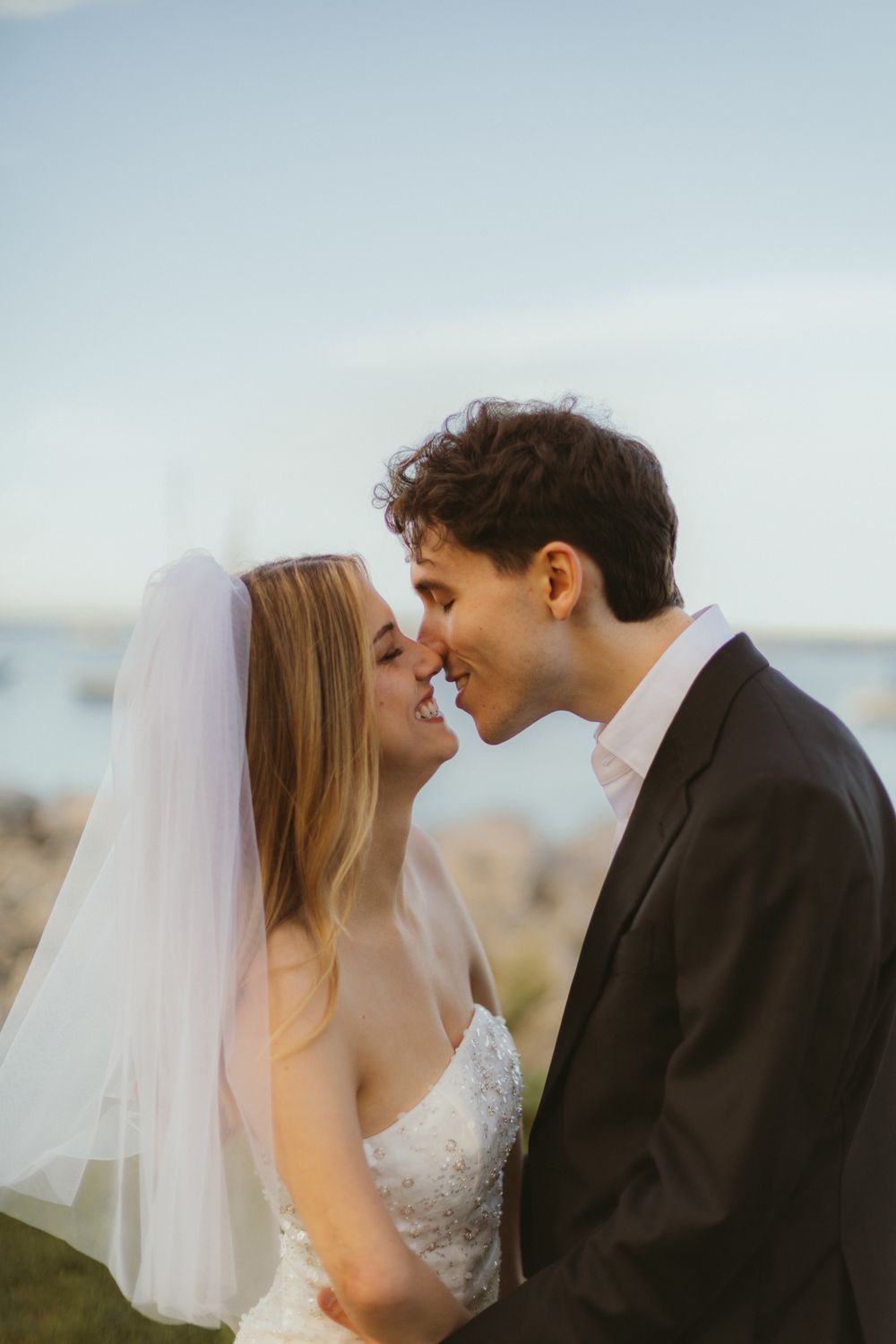 Bride in a veil and beaded gown leaning in to kiss her groom at golden hour, calm harbor water behind them