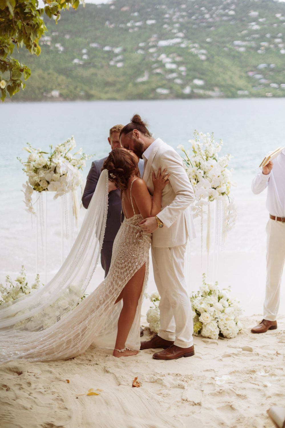 Bride laughing during beach vows with bridesmaids in brown dresses and a white floral arch, turquoise water in the background