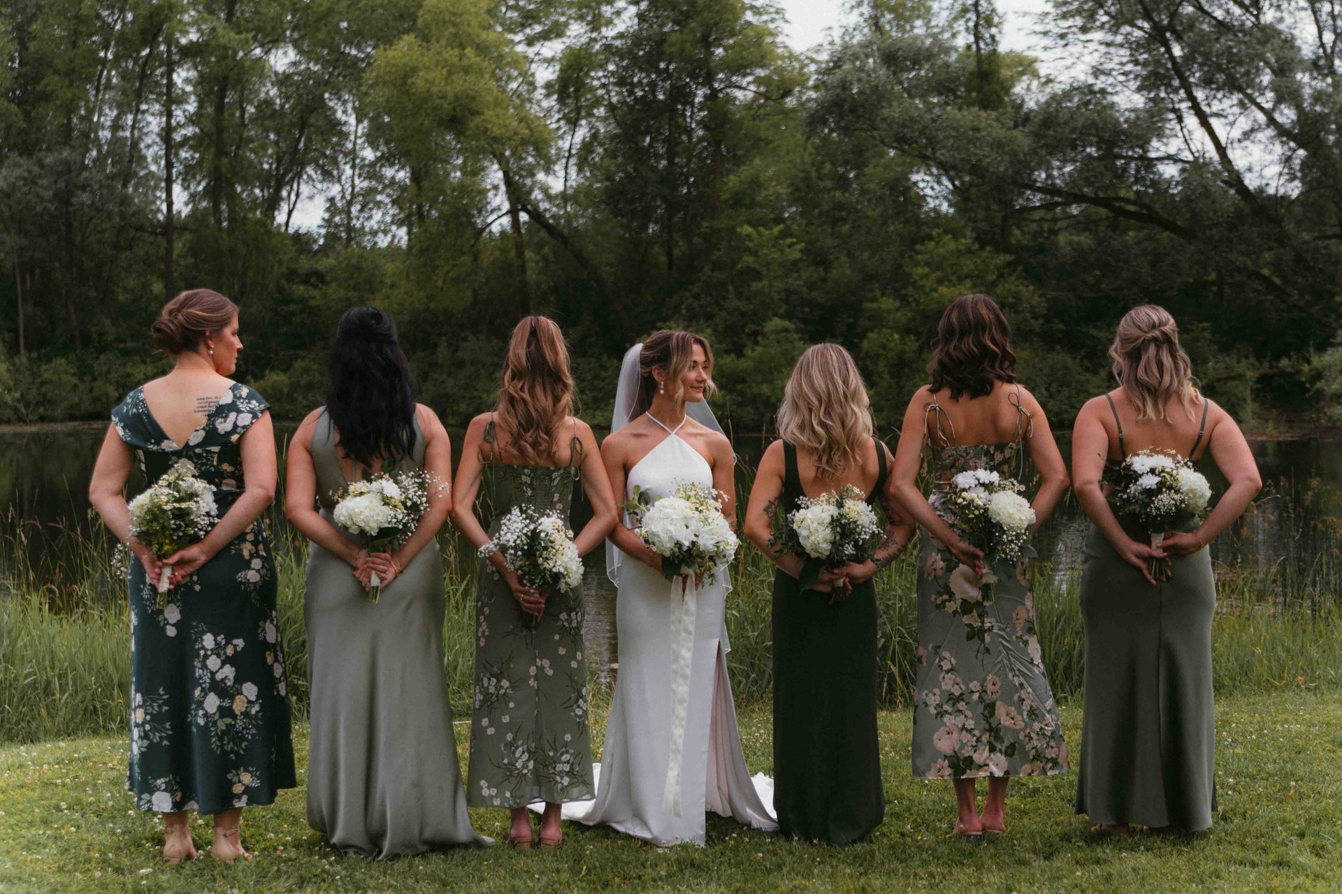 Bride and bridesmaids standing in a row holding bouquets, backs to the camera, overlooking a pond