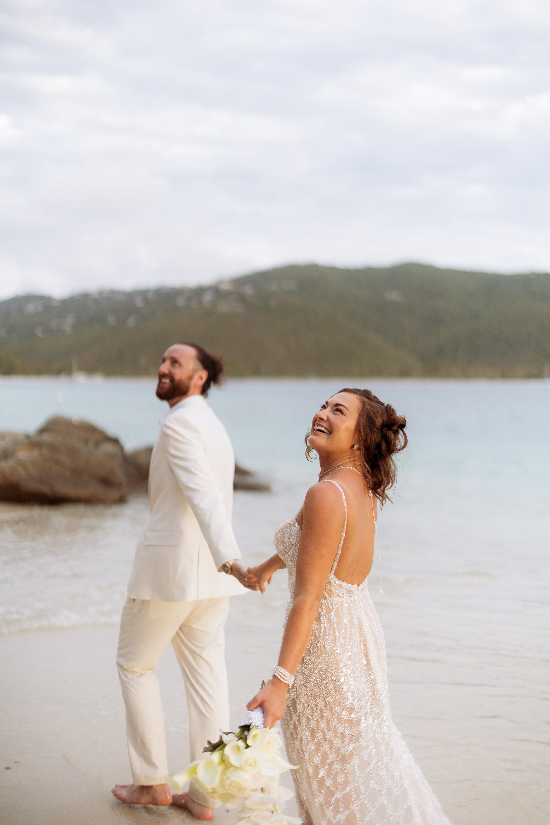 Laughing bride glancing back as groom leads her along a sandy beach, turquoise water and green hills behind them