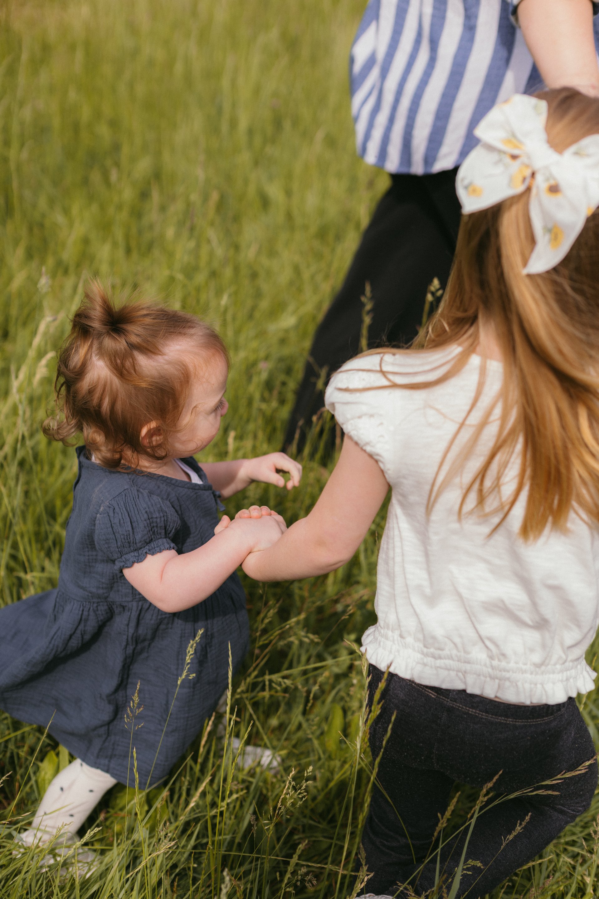 Young girl in a white bow holding hands with a toddler in a blue dress in a sunlit grass field