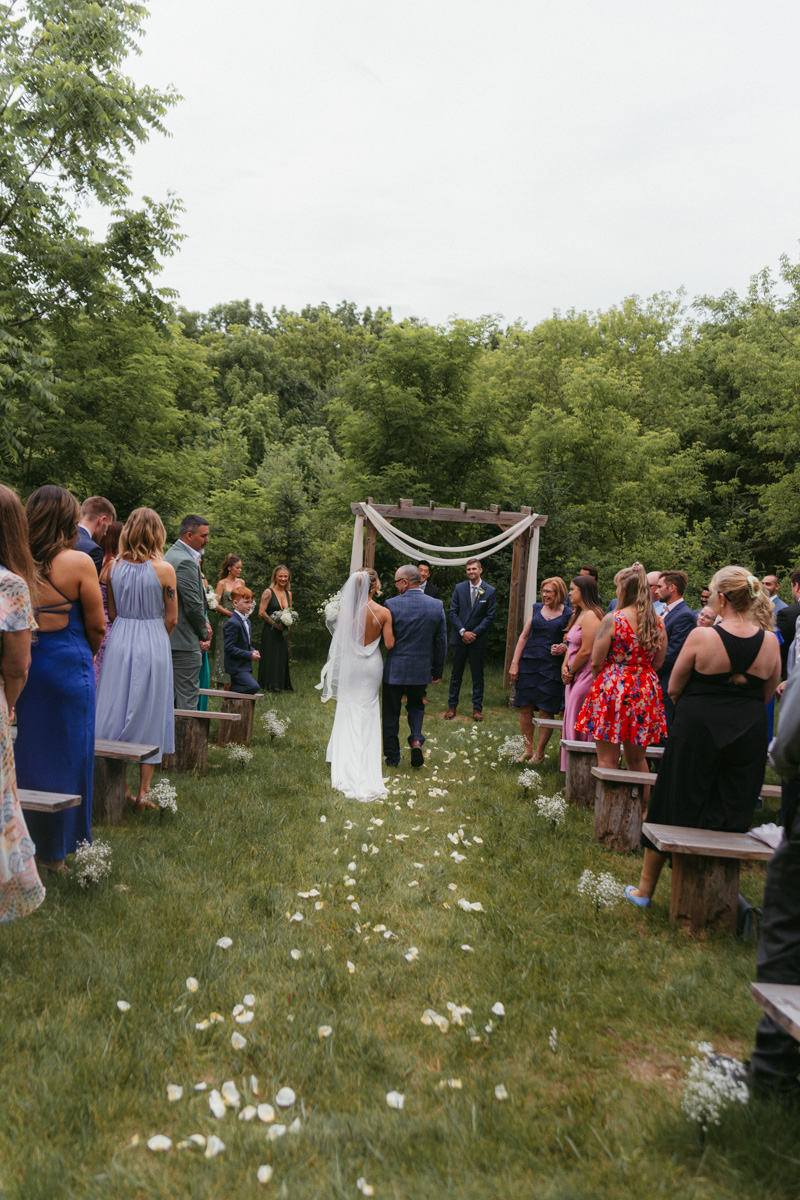 Bride walking down a flower-petal aisle escorted by her father, guests standing on either side, rustic wooden arch ahead