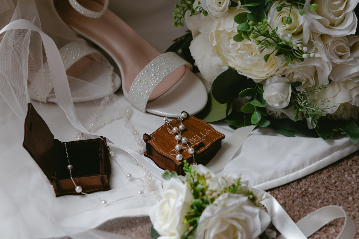 Flat lay of bridal details — crystal heels, white rose bouquet, pearl earrings in a wooden jewelry box, and a veil