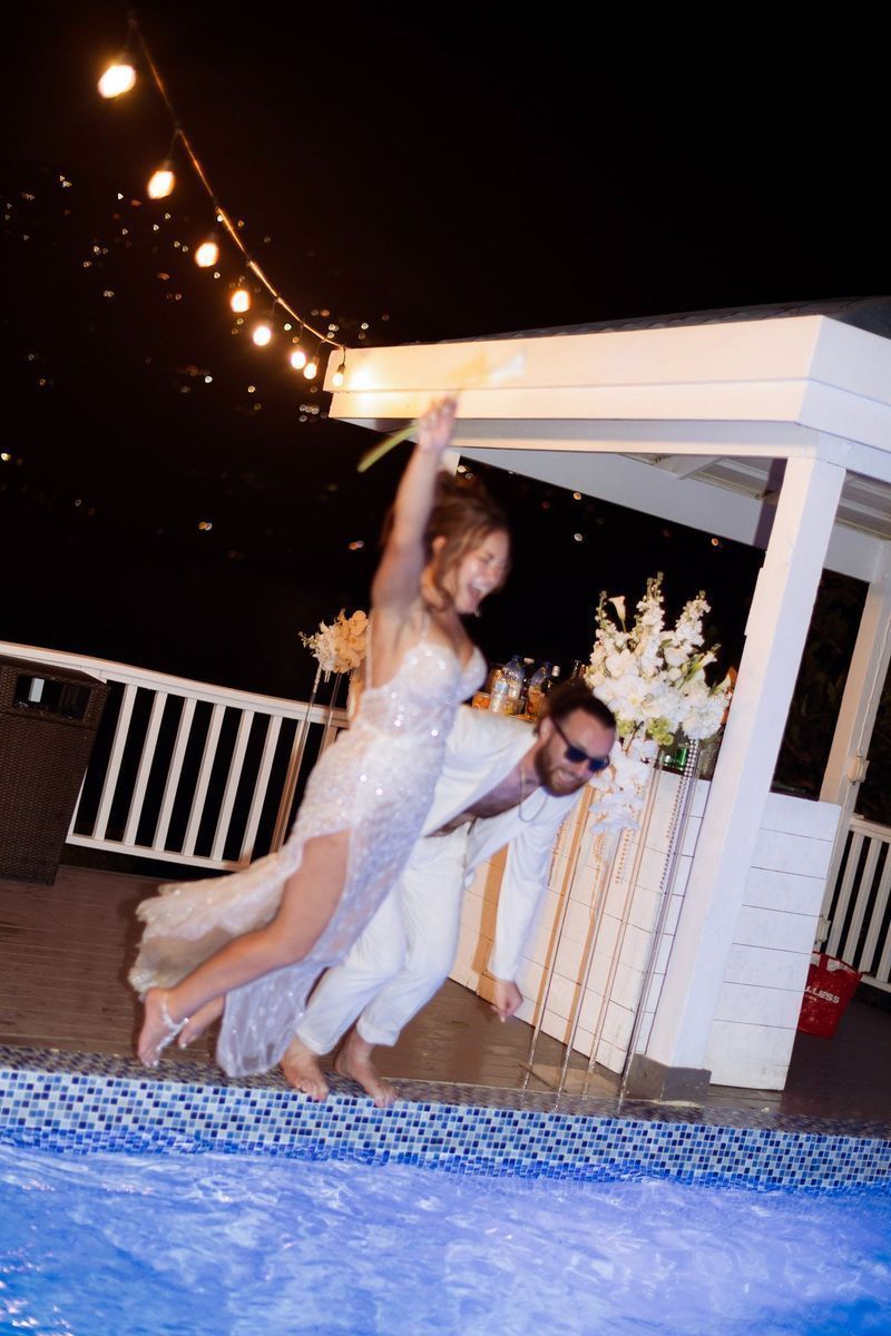 Bride laughing as groom lifts her near a rooftop pool at night, string lights above